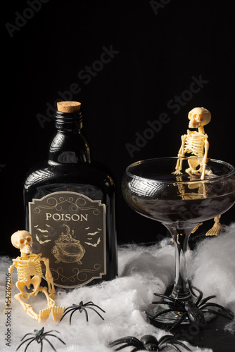 Top view of black Halloween cocktail glass with skeletons and bottle of poison on spider web, black background, vertical