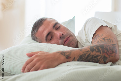Handsome man with tattoo on his arm sleeping on pillow in bed. Medium shot of Caucasian man relaxing on weekend at home, having rest after working day. Sleep cycle, dream concept