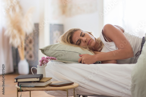 Carefree Caucasian woman sleeping on her side in bed in morning. Medium shot of beautiful woman in pajamas relaxing in cozy bedroom with bedside table. Dream, sleeping concept
