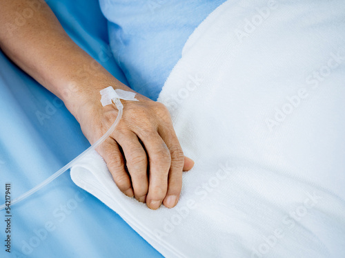 Close up saline solution in senior patients hand in blue gown lying on the bed with white blanket in hospital room with copy space. Elder patient's hand receiving iv saline solution.
