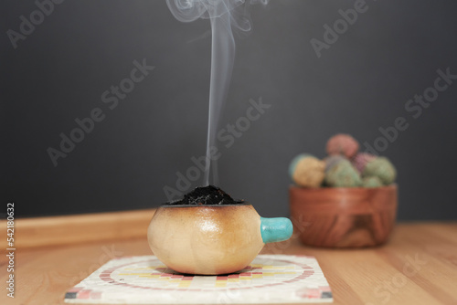 Smoke rising from a ceramic bowl on a wooden table, mosaic plate and black background.