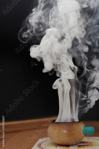 Smoke rising from a ceramic bowl on a wooden table, mosaic plate and black background.