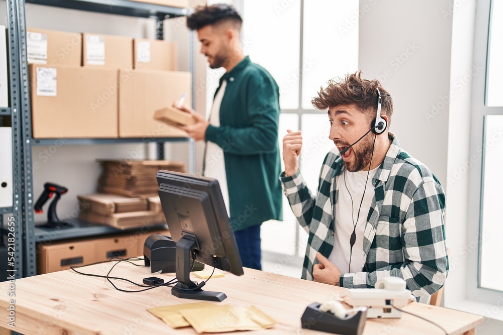 Two hispanic men working at small business ecommerce pointing thumb up to the side smiling happy with open mouth Two hispanic men working at small business ecommerce pointing thumb up to the side smiling happy with open mouth