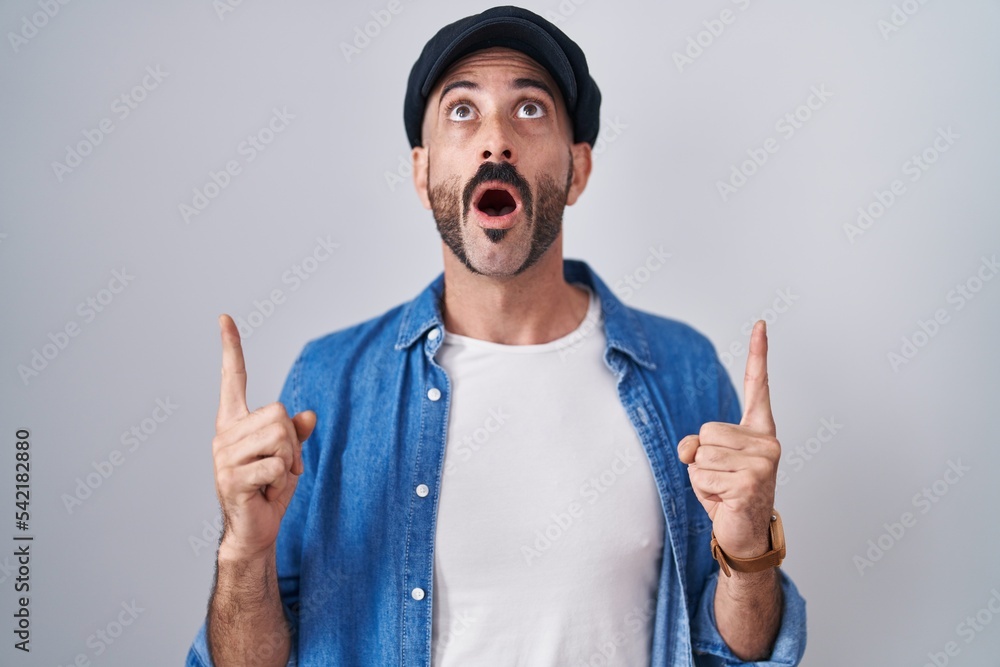 Hispanic man with beard standing over isolated background amazed and surprised looking up and pointing with fingers and raised arms. Hispanic man with beard standing over isolated background amazed and surprised looking up and pointing with fingers and raised arms.