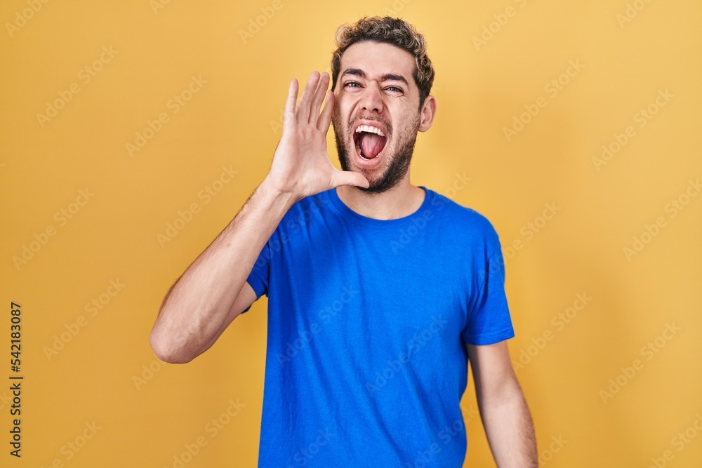 Hispanic man with beard standing over yellow background shouting and screaming loud to side with hand on mouth. communication concept. Hispanic man with beard standing over yellow background shouting and screaming loud to side with hand on mouth. communication concept.