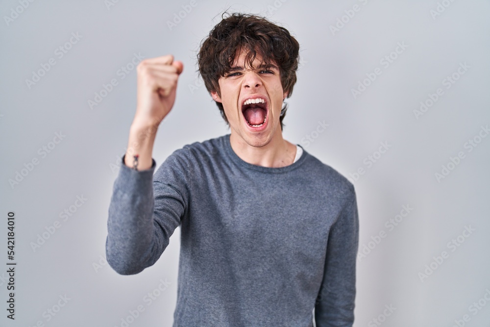 Young man standing over isolated background angry and mad raising fist frustrated and furious while shouting with anger. rage and aggressive concept. Young man standing over isolated background angry and mad raising fist frustrated and furious while shouting with anger. rage and aggressive concept.