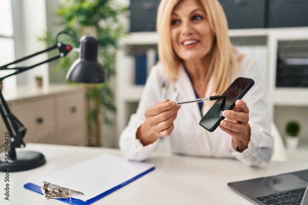 Middle age blonde woman wearing doctor uniform showing screen smartphone at clinic Middle age blonde woman wearing doctor uniform showing screen smartphone at clinic