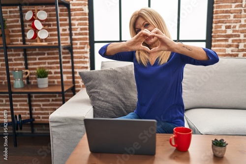 Middle age blonde woman using computer laptop at home smiling in love doing heart symbol shape with hands. romantic concept.