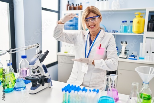 Middle age blonde woman working at scientist laboratory gesturing with hands showing big and large size sign  measure symbol. smiling looking at the camera. measuring concept.