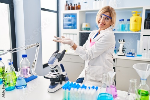 Middle age blonde woman working at scientist laboratory pointing aside with hands open palms showing copy space  presenting advertisement smiling excited happy