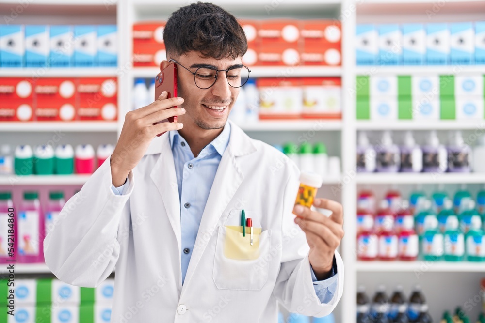 Young hispanic man pharmacist holding pills bottle talking on smartphone at pharmacy Young hispanic man pharmacist holding pills bottle talking on smartphone at pharmacy