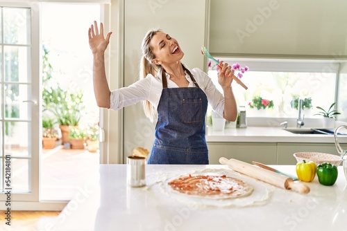 Young blonde woman singing song using spoon as a microphone cooking pizza at kitchen