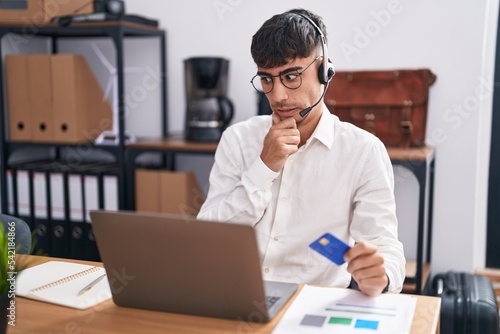 Young hispanic man working using computer laptop holding credit card thinking worried about a question, concerned and nervous with hand on chin