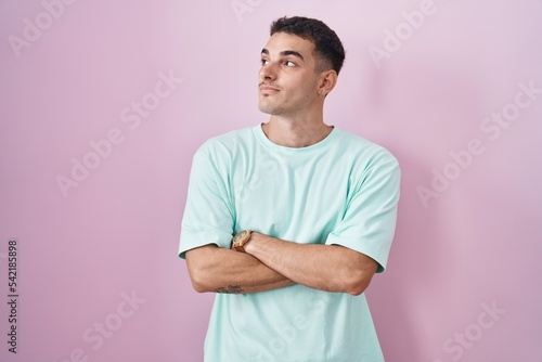 Handsome hispanic man standing over pink background looking to the side with arms crossed convinced and confident