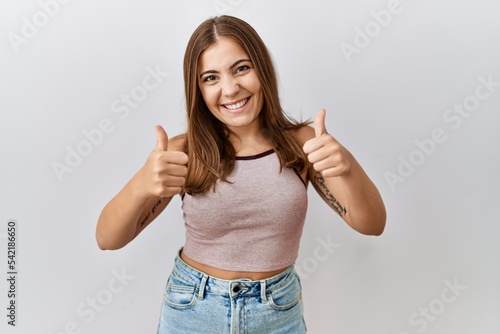 Young brunette woman standing over isolated background success sign doing positive gesture with hand, thumbs up smiling and happy. cheerful expression and winner gesture. © Krakenimages.com