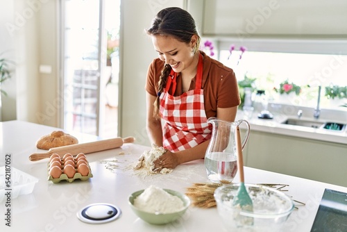 Young beautiful hispanic woman smiling confident kneading bread dough with hands at the kitchen