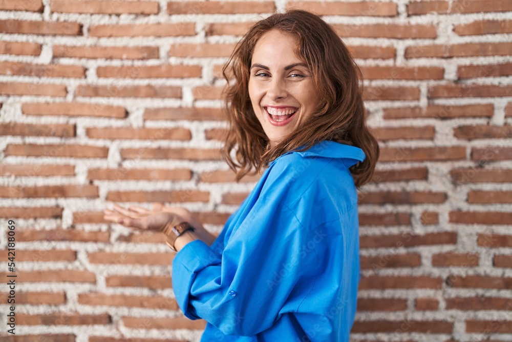 Beautiful brunette woman standing over bricks wall pointing aside with hands open palms showing copy space, presenting advertisement smiling excited happy