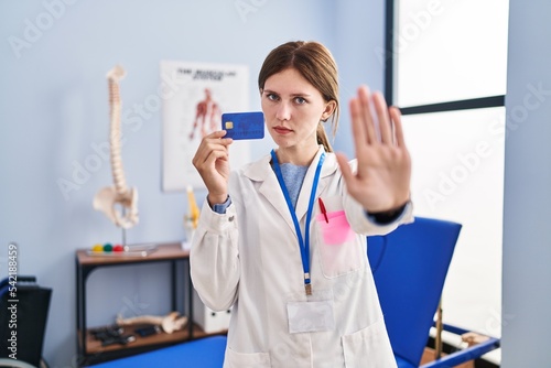 Young brunette woman working at pain recovery clinic holding credit card with open hand doing stop sign with serious and confident expression  defense gesture