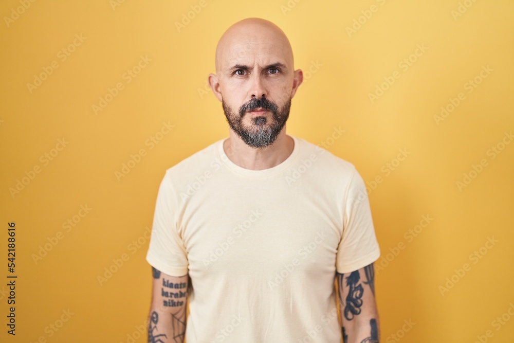 Hispanic man with tattoos standing over yellow background relaxed with serious expression on face. simple and natural looking at the camera. Hispanic man with tattoos standing over yellow background relaxed with serious expression on face. simple and natural looking at the camera.