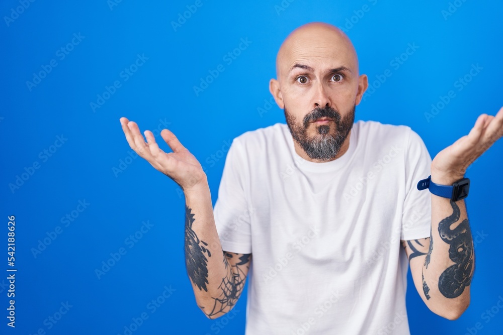 Naklejka premium Hispanic man with tattoos standing over blue background clueless and confused expression with arms and hands raised. doubt concept.