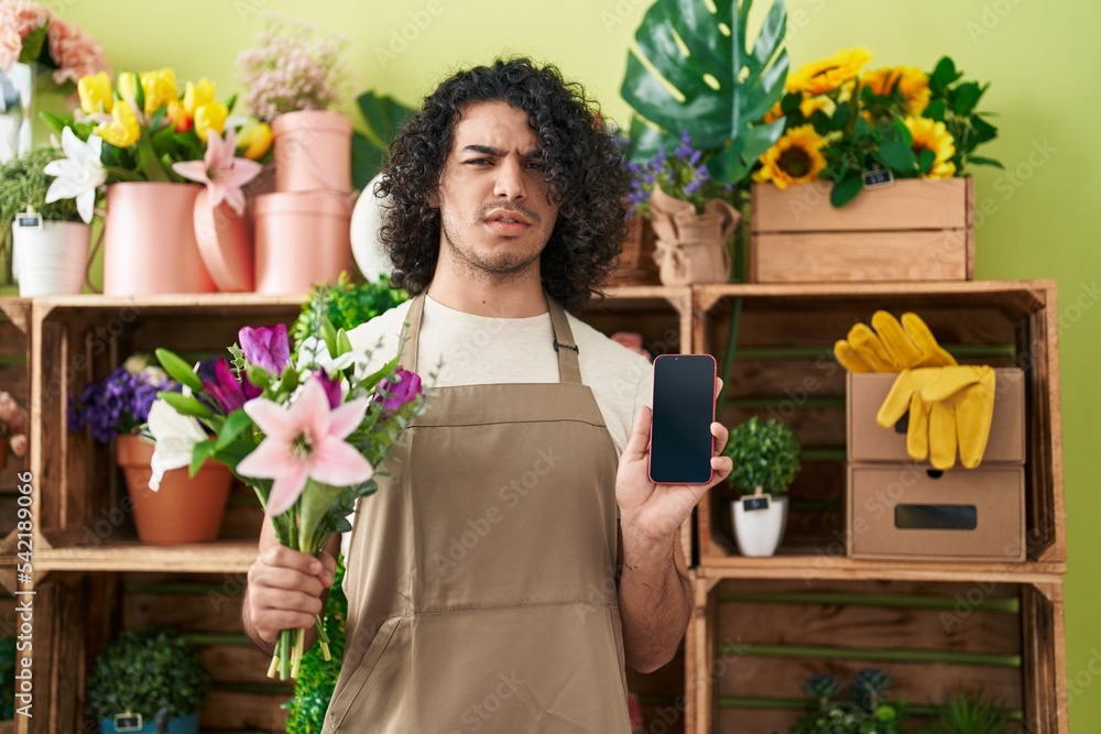 Hispanic man with curly hair working at florist shop showing smartphone screen clueless and confused expression. doubt concept.