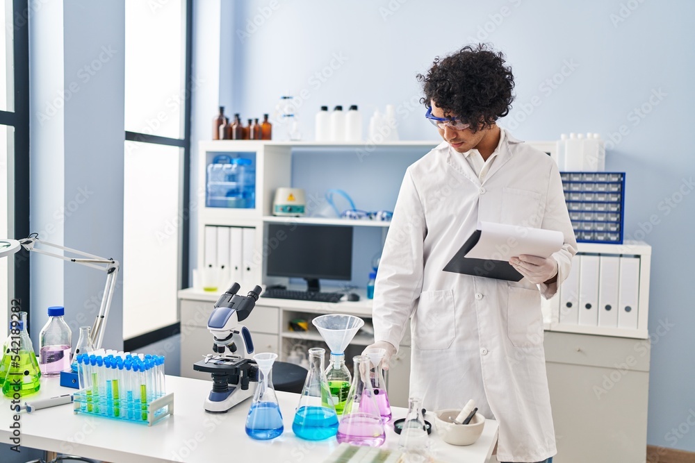Young hispanic man wearing scientist uniform holding clipboard and test tube at laboratory Young hispanic man wearing scientist uniform holding clipboard and test tube at laboratory