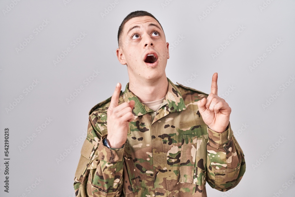 Young man wearing camouflage army uniform amazed and surprised looking up and pointing with fingers and raised arms. Young man wearing camouflage army uniform amazed and surprised looking up and pointing with fingers and raised arms.