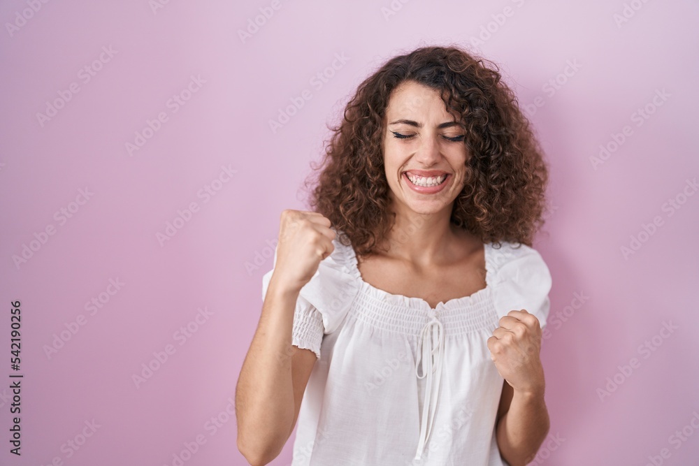 Hispanic woman with curly hair standing over pink background very happy and excited doing winner gesture with arms raised, smiling and screaming for success. celebration concept. Hispanic woman with curly hair standing over pink background very happy and excited doing winner gesture with arms raised, smiling and screaming for success. celebration concept.