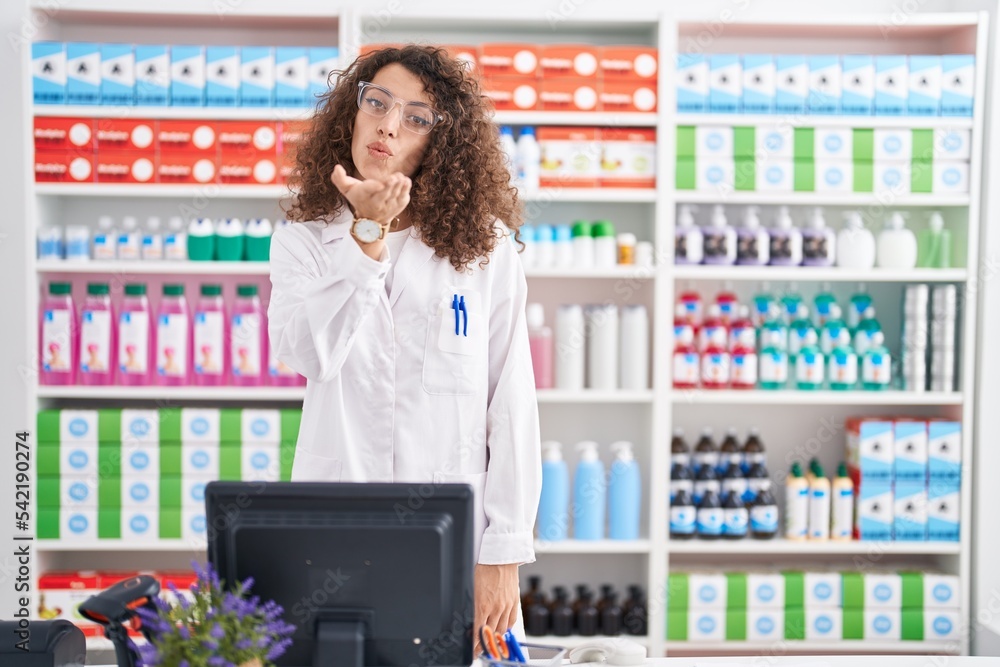 Hispanic woman with curly hair working at pharmacy drugstore looking at the camera blowing a kiss with hand on air being lovely and sexy. love expression.