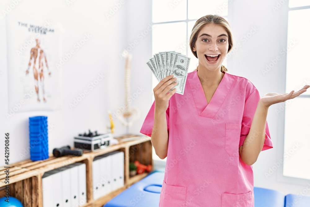 Young physiotherapist woman working at pain recovery clinic holding dollars celebrating achievement with happy smile and winner expression with raised hand