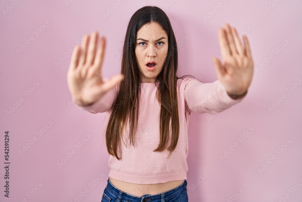 Young brunette woman standing over pink background doing stop gesture with hands palms, angry and frustration expression Young brunette woman standing over pink background doing stop gesture with hands palms, angry and frustration expression