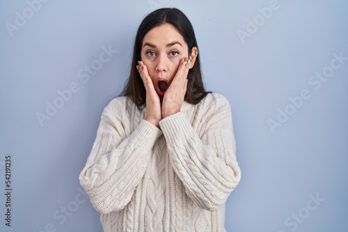 Young brunette woman standing over blue background afraid and shocked, surprise and amazed expression with hands on face