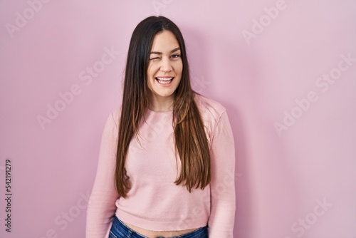 Young brunette woman standing over pink background winking looking at the camera with sexy expression, cheerful and happy face.