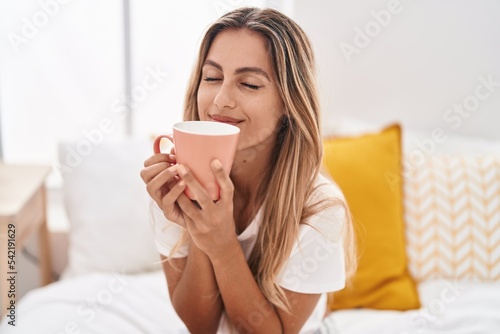 Young blonde woman drinking cup of coffee sitting on bed at bedroom