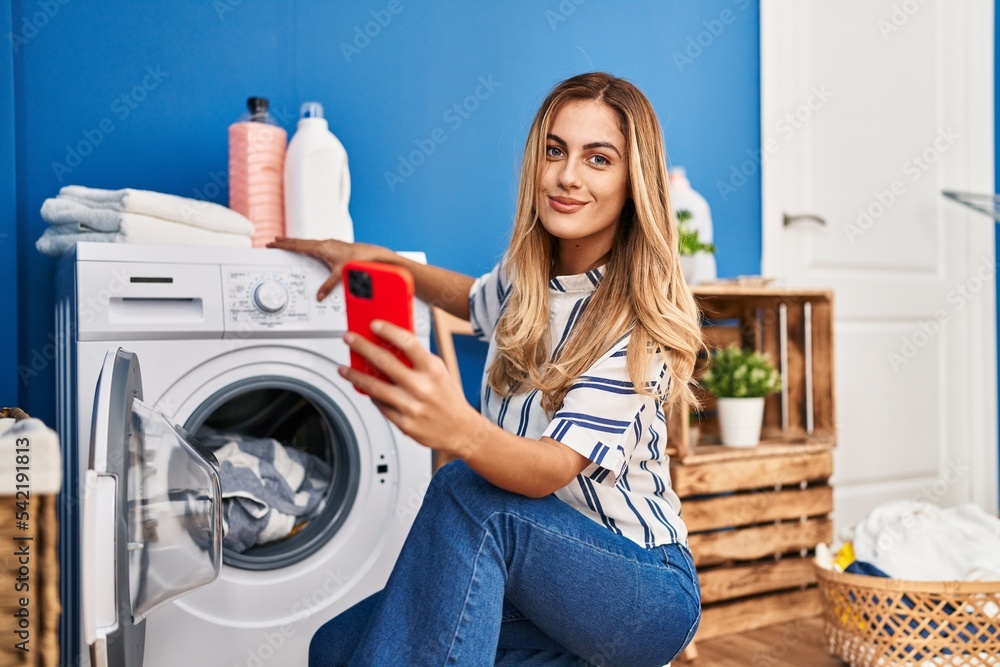 Young blonde woman using smartphone and washing clothes at laundry room Young blonde woman using smartphone and washing clothes at laundry room