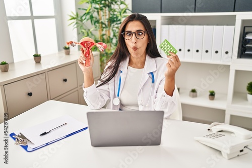 Young hispanic doctor woman holding anatomical female genital organ and birth control pills making fish face with mouth and squinting eyes, crazy and comical. photo