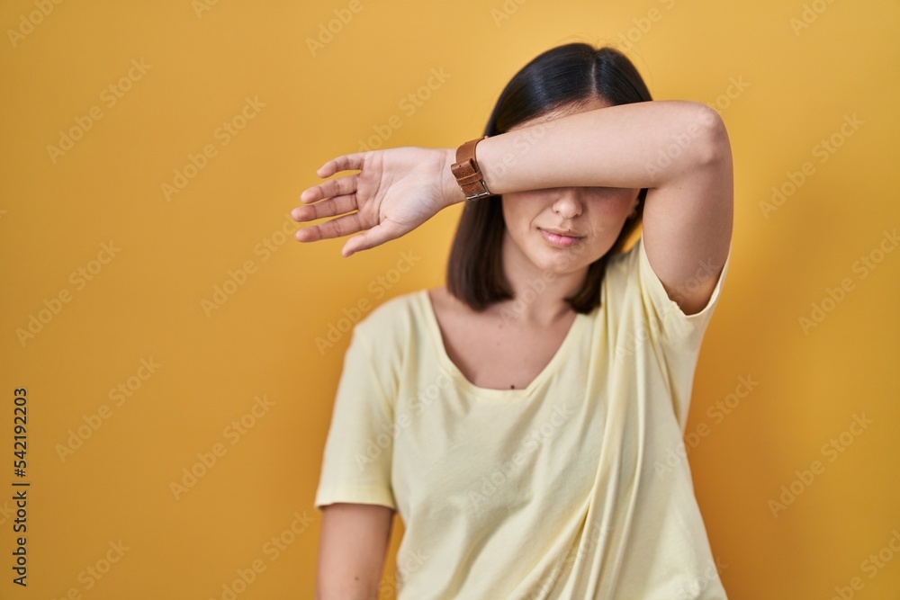Hispanic girl wearing casual t shirt over yellow background covering eyes with arm, looking serious and sad. sightless, hiding and rejection concept Hispanic girl wearing casual t shirt over yellow background covering eyes with arm, looking serious and sad. sightless, hiding and rejection concept