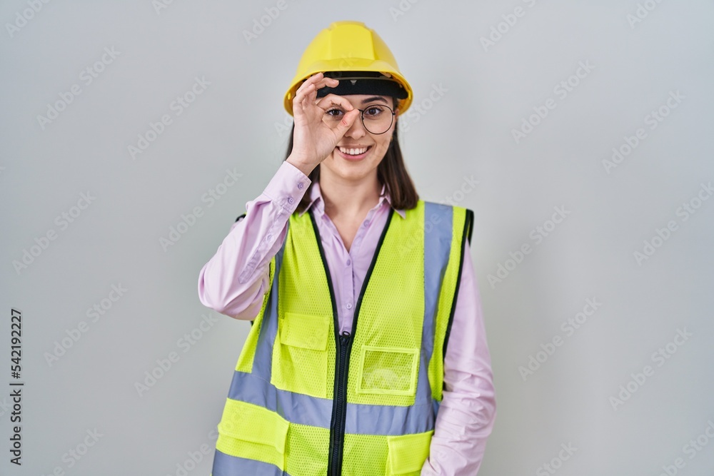 Hispanic girl wearing builder uniform and hardhat doing ok gesture with hand smiling, eye looking through fingers with happy face.