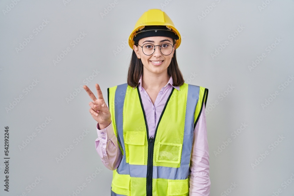 Hispanic girl wearing builder uniform and hardhat smiling looking to the camera showing fingers doing victory sign. number two. Hispanic girl wearing builder uniform and hardhat smiling looking to the camera showing fingers doing victory sign. number two.