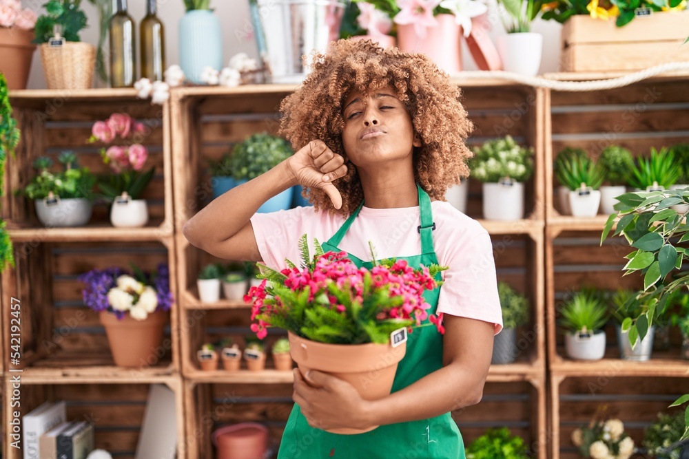 Young hispanic woman with curly hair working at florist shop holding plant with angry face, negative sign showing dislike with thumbs down, rejection concept