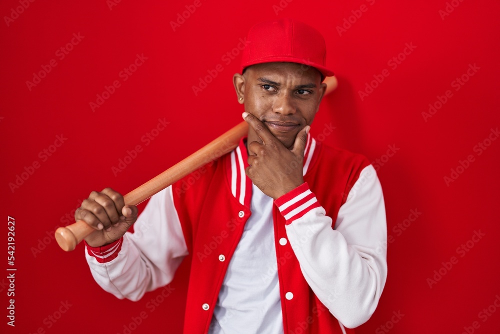 Young hispanic man playing baseball holding bat with hand on chin thinking about question, pensive expression. smiling and thoughtful face. doubt concept. Young hispanic man playing baseball holding bat with hand on chin thinking about question, pensive expression. smiling and thoughtful face. doubt concept.
