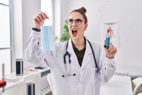 Young hispanic girl holding medical asthma inhaler at the clinic angry and mad screaming frustrated and furious, shouting with anger looking up.