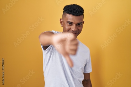 Young hispanic man standing over yellow background looking unhappy and angry showing rejection and negative with thumbs down gesture. bad expression.