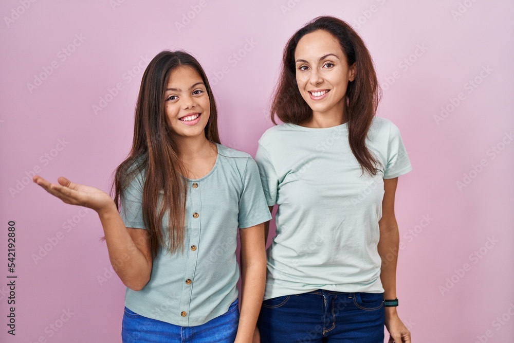 Young mother and daughter standing over pink background smiling cheerful presenting and pointing with palm of hand looking at the camera. Young mother and daughter standing over pink background smiling cheerful presenting and pointing with palm of hand looking at the camera.