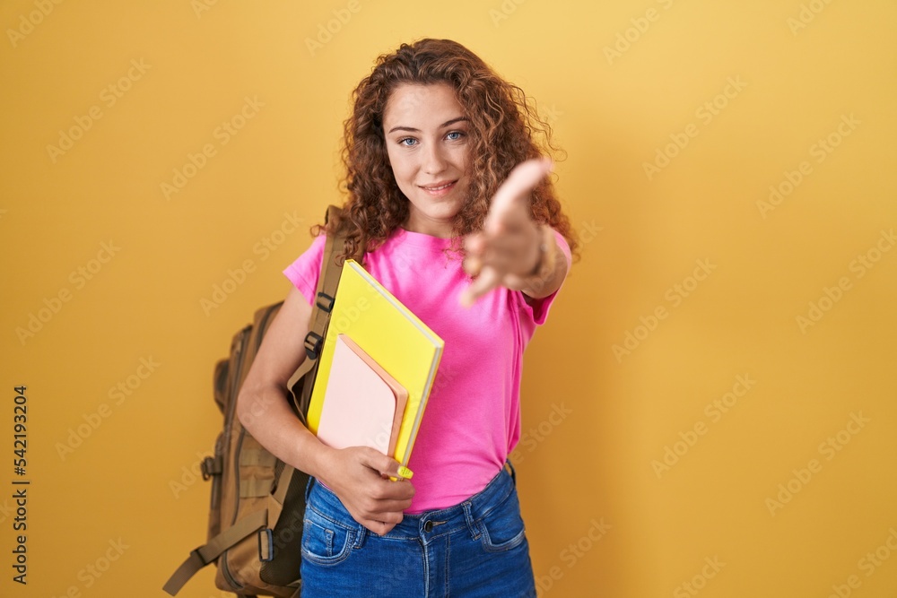 Young caucasian woman wearing student backpack and holding books smiling friendly offering handshake as greeting and welcoming. successful business. Young caucasian woman wearing student backpack and holding books smiling friendly offering handshake as greeting and welcoming. successful business.