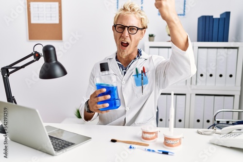 Young blond dentist man holding mouthwash at clinic annoyed and frustrated shouting with anger, yelling crazy with anger and hand raised