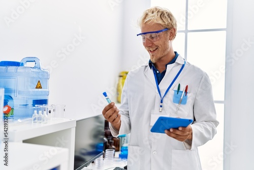 Young caucasian man wearing scientist uniform holding test tube and touchpad at laboratory