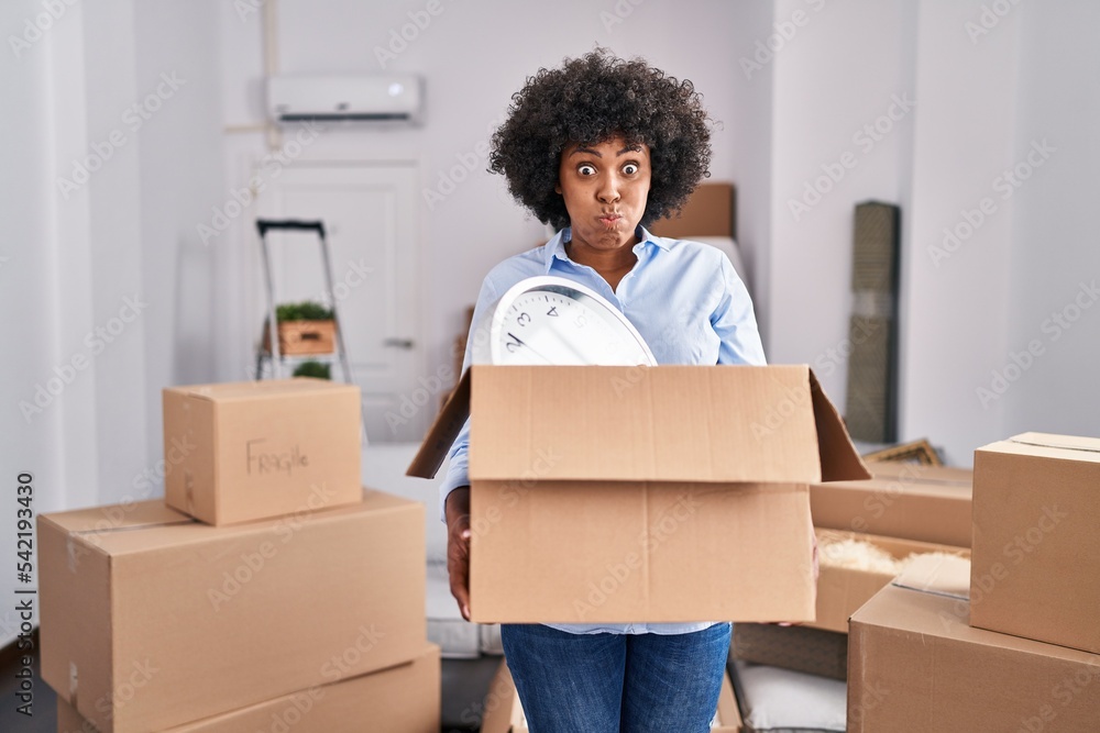 Black woman with curly hair moving to a new home holding cardboard box puffing cheeks with funny face. mouth inflated with air, catching air. - obrazy, fototapety, plakaty 