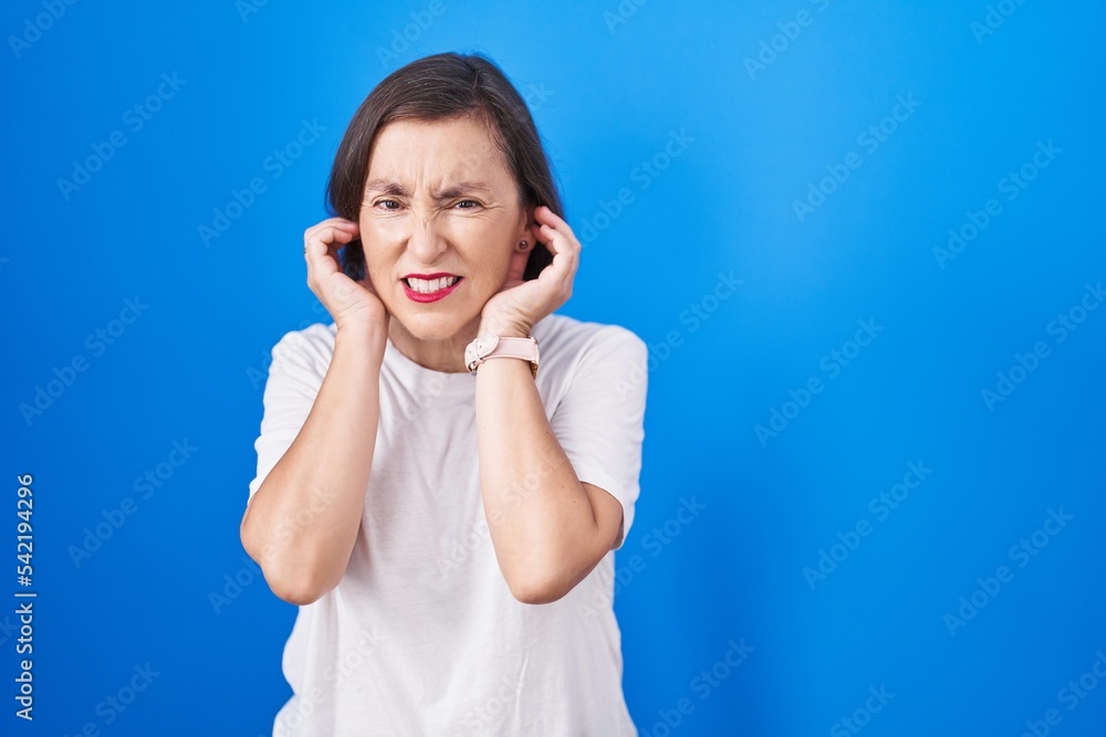 Middle age hispanic woman standing over blue background covering ears with fingers with annoyed expression for the noise of loud music. deaf concept. Middle age hispanic woman standing over blue background covering ears with fingers with annoyed expression for the noise of loud music. deaf concept.
