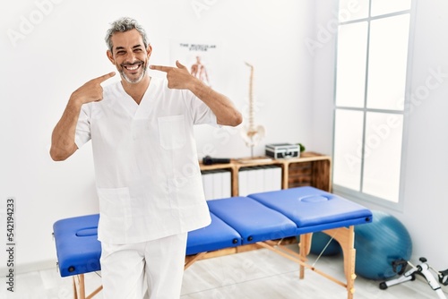 Middle age hispanic therapist man working at pain recovery clinic smiling cheerful showing and pointing with fingers teeth and mouth. dental health concept.